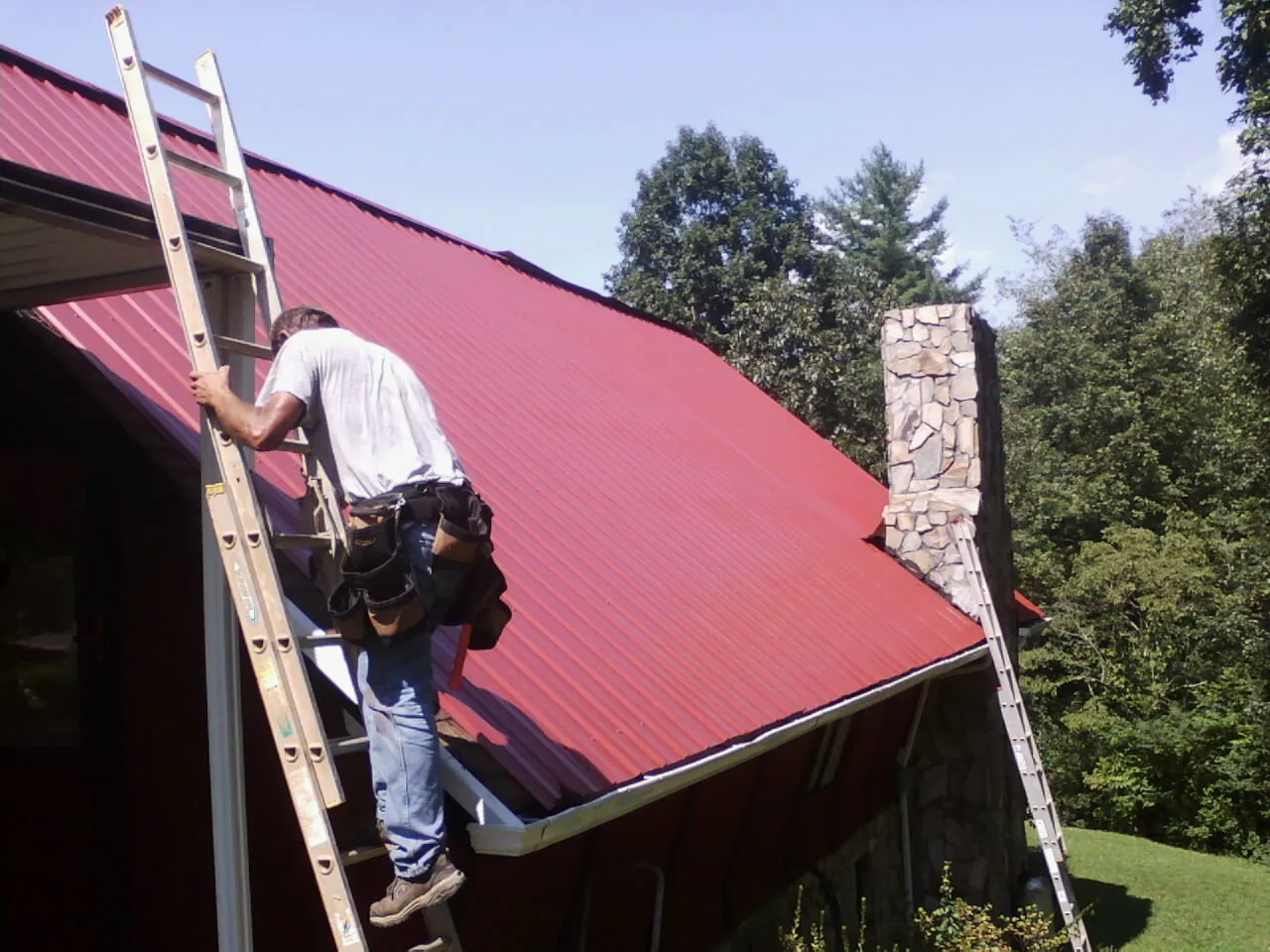 Roofer on a ladder beside a red metal roof and stone chimney — Soffit & Fascia Repair by Roofing Brothers in Tarrs